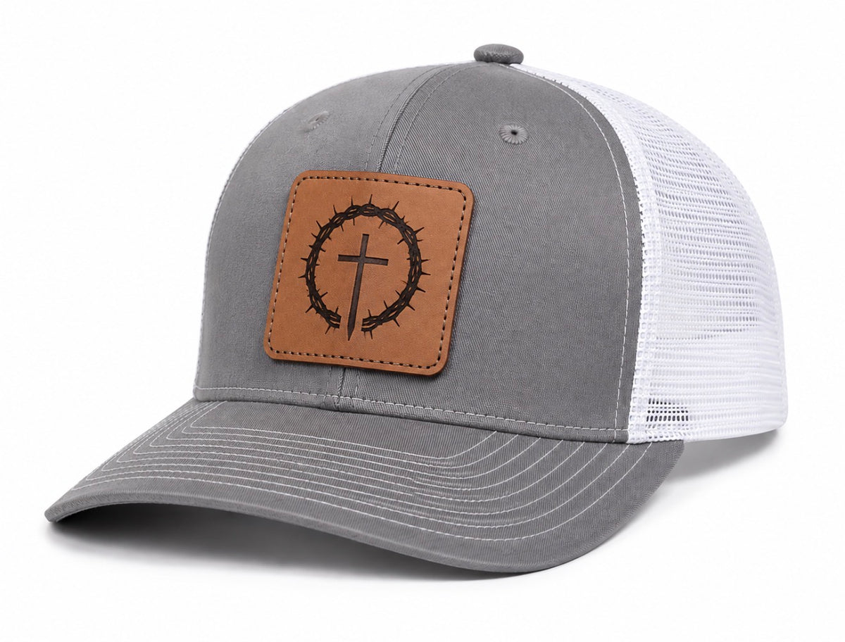 A gray and white trucker hat with a brown faux-leather crown patch featuring a black thorn crown and cross design, shown on a white background.